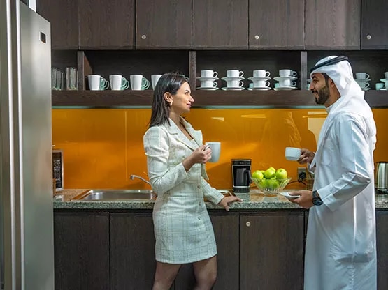 Two professionals having a coffee chat in the modern kitchen area at Sahab Tower, Kuwait, featuring sleek cabinetry and a fully equipped setup.